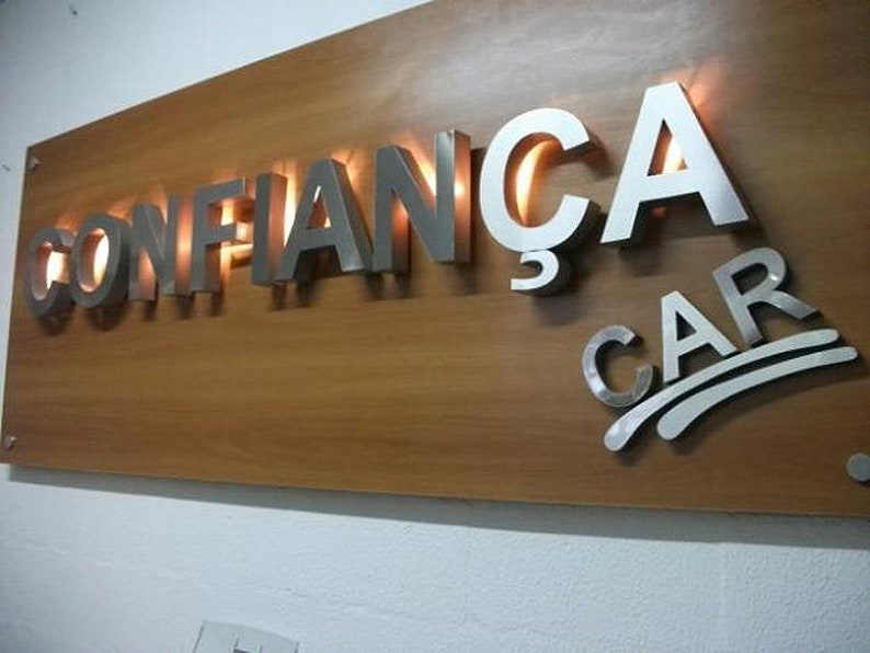 May include: A wooden sign with the words "CONFIAN&Ccedil;A CAR" in raised, silver letters. The letters are illuminated from behind, creating a warm glow.