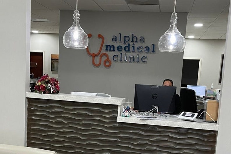 May include: A reception desk with a wavy dark brown front panel and a light-colored countertop. On the gray wall behind the desk, a sign reads alpha medical clinic in dark letters with an orange stethoscope graphic. Two glass pendant lights hang above.