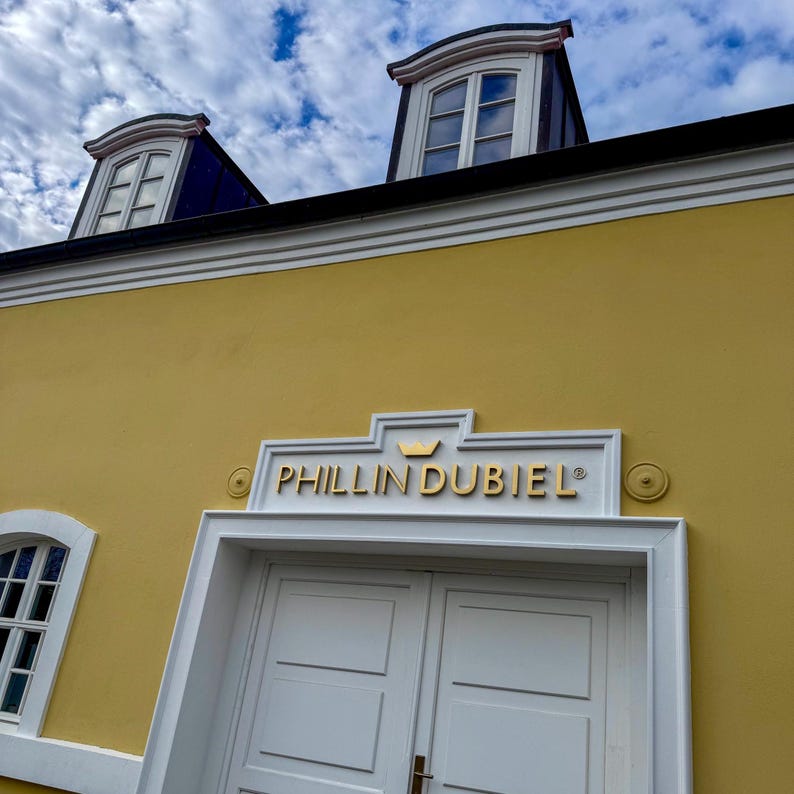 Puede incluir: Una fachada de edificio amarilla con una entrada de doble puerta blanca y un letrero encima que dice PHILLIN DUBIEL con un peque&ntilde;o icono de corona. La estructura incluye molduras blancas, dos ventanas arqueadas en el techo y cielo azul.