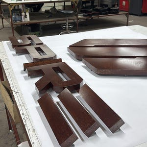May include: A set of three-dimensional, dark brown, glossy letters spelling the word "SPRI" arranged on a white surface. The letters have a thick, block-like construction with sharp, clean edges and a smooth, reflective finish, positioned in a workshop.