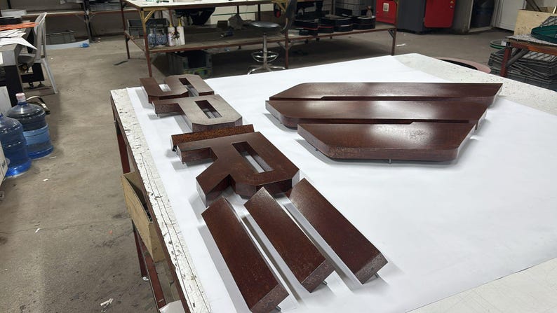 May include: A set of three-dimensional, dark brown, glossy letters spelling the word "SPRI" arranged on a white surface. The letters have a thick, block-like construction with sharp, clean edges and a smooth, reflective finish, positioned in a workshop.