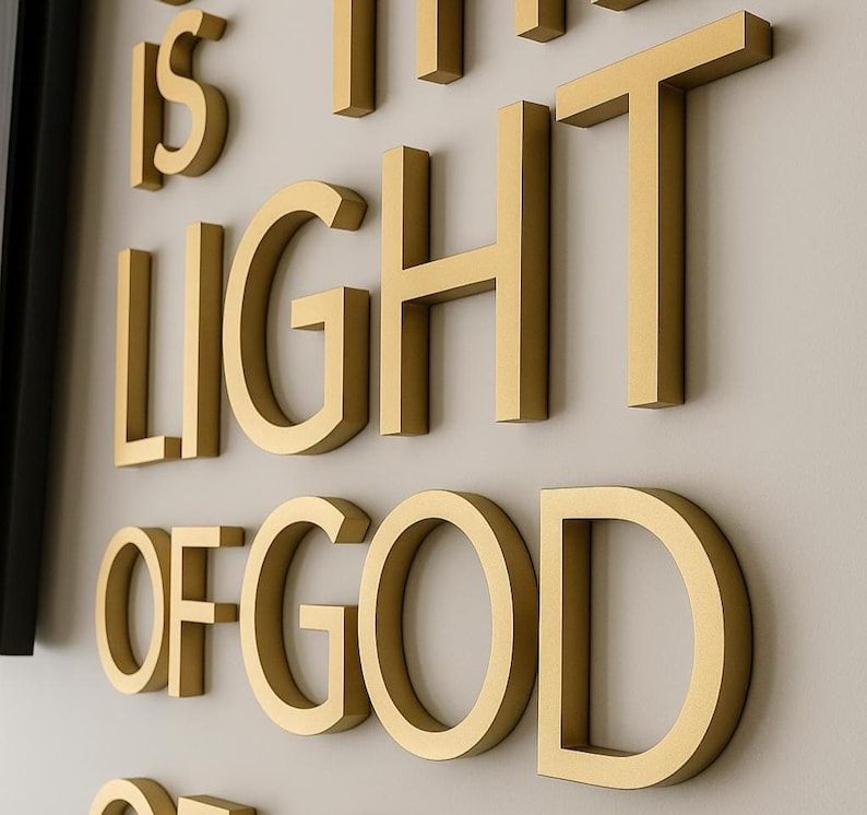K&ouml;nnte beinhalten: Goldfarbene, dreidimensionale Buchstaben schreiben "IS THE LIGHT OF GOD" an eine hellfarbene Wand. Die Buchstaben sind dick und haben ein metallisches Aussehen, wodurch ein dekoratives Wandkunstwerk entsteht.