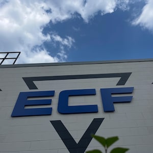 Puede incluir: Una pared de bloques de hormig&oacute;n claro presenta un letrero grande y llamativo de color azul oscuro con las letras ECF en una fuente sans-serif. Encima y debajo de las letras hay formas geom&eacute;tricas triangulares negras bajo un cielo azul.