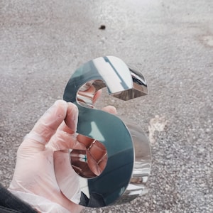 K&ouml;nnte beinhalten: Ein gl&auml;nzender, verchromter Buchstabe "S", gehalten in einer behandschuhten Hand. Die Metalloberfl&auml;che spiegelt die Umgebung wider. Der Buchstabe hat ein k&uuml;hnes, stilisiertes Design, wahrscheinlich f&uuml;r Beschilderungen oder dekorative Zwecke.