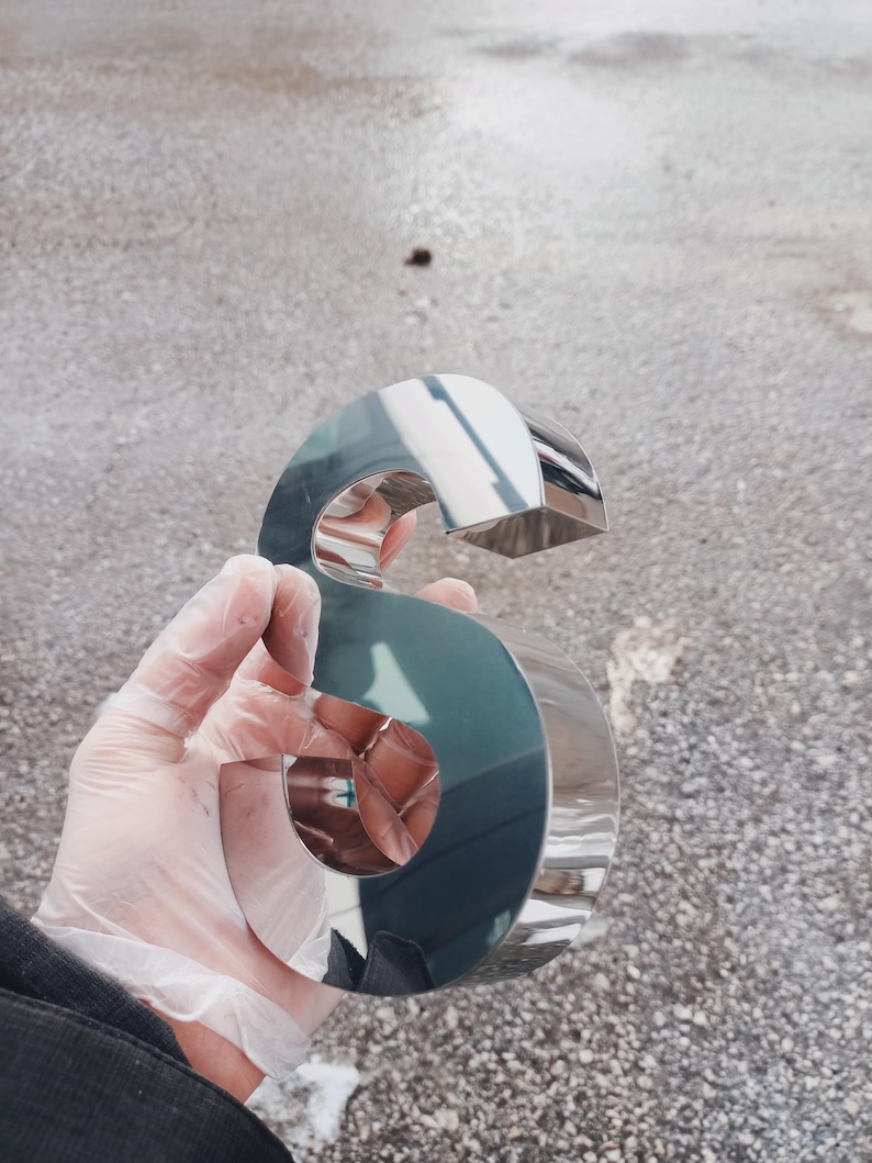 K&ouml;nnte beinhalten: Ein gl&auml;nzender, verchromter Buchstabe "S", gehalten in einer behandschuhten Hand. Die Metalloberfl&auml;che spiegelt die Umgebung wider. Der Buchstabe hat ein k&uuml;hnes, stilisiertes Design, wahrscheinlich f&uuml;r Beschilderungen oder dekorative Zwecke.