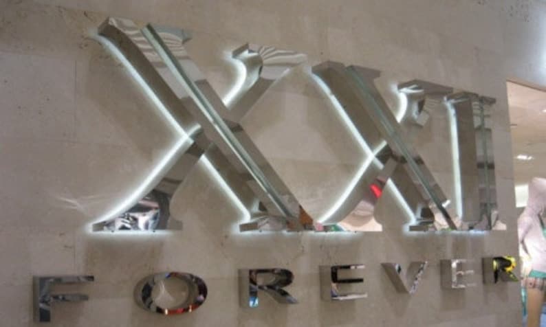 May include: A silver metal sign with the words "XXI FOREVER" in large, bold letters. The letters are illuminated with white lights.