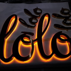 May include: A black metal sign with the word "lolo" in a cursive font. The sign is illuminated with warm white LED lights.