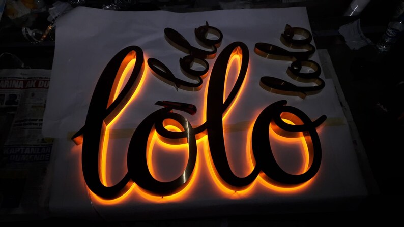 May include: A black metal sign with the word "lolo" in a cursive font. The sign is illuminated with warm white LED lights.