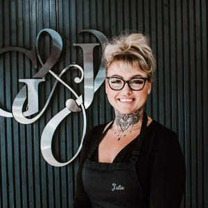 May include: A person with blonde hair in a messy bun, wearing black-rimmed glasses and a black apron with the name Julie embroidered in white script, standing in front of a wall featuring large, metallic, stylized letters G and J in a cursive font.