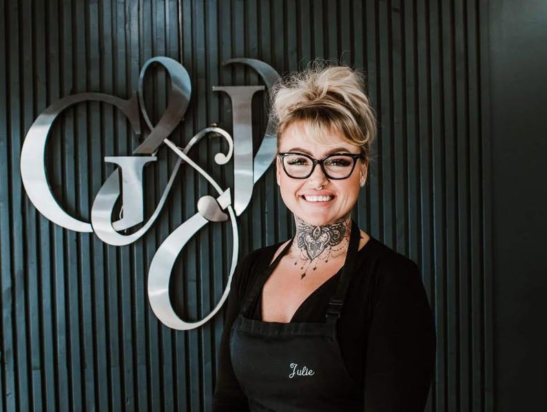 May include: A person with blonde hair in a messy bun, wearing black-rimmed glasses and a black apron with the name Julie embroidered in white script, standing in front of a wall featuring large, metallic, stylized letters G and J in a cursive font.