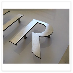 May include: Close-up of a silver metal letter "R" with a modern font, mounted on a white surface. The letter has a brushed metal finish and is elevated slightly above the surface by small posts.