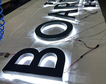 以下が含まれることがあります： 「BOARDWALK HOME」と書かれた黒い照明付きチャンネル文字。文字は白い表面に取り付けられ、明るい白色LEDバックライトが付いています。文字はワイヤーで接続されており、組み立て中であることを示唆しています。