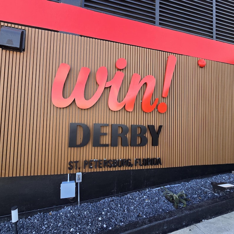Puede incluir: Un exterior de edificio con una pared de listones de madera verticales y letreros tridimensionales. La palabra win! est&aacute; en letra cursiva roja grande, seguida de la palabra DERBY en letras negras y St. Petersburg, Florida en texto negro.