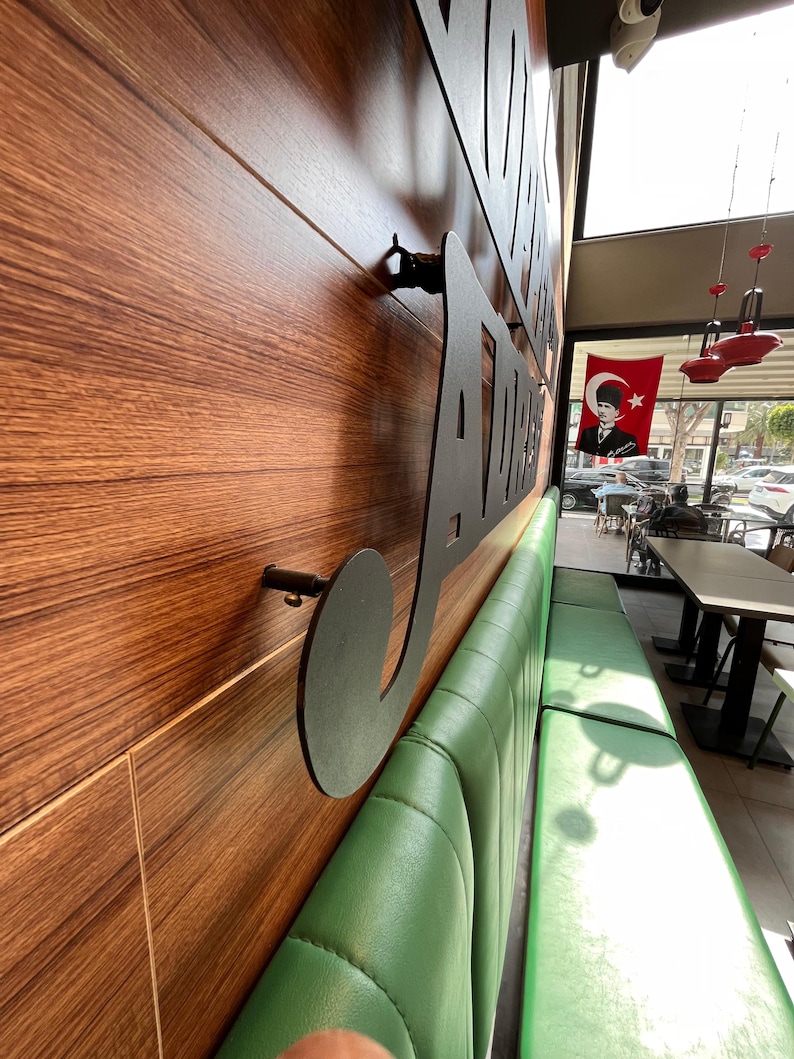 May include: Interior shot of a restaurant with a wooden wall featuring a large, black metal sign. A long, green leather bench runs along the wall. A Turkish flag hangs in the background.