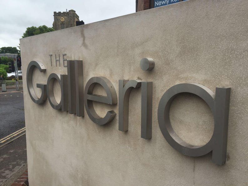 May include: A beige concrete wall featuring the three-dimensional metallic silver text The Galleria. The letters are mounted to the surface, with a small circular dot above the letter i. The sign is positioned outdoors in an urban setting.