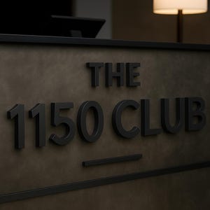 May include: A dark gray rectangular sign featuring the raised black text THE 1150 CLUB in a bold sans-serif font. The sign has a matte finish with a thin black border along the top and bottom edges, and a horizontal black line centered below the text.