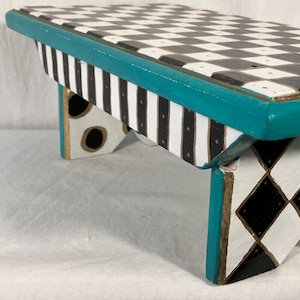 May include: A small wooden stool featuring a black and white checkered top, a teal painted edge, and sides decorated with black and white vertical stripes, black dots, and a black and white diamond pattern on the legs with gold-toned accents.
