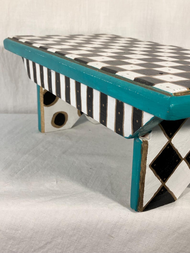 May include: A small wooden stool featuring a black and white checkered top, a teal painted edge, and sides decorated with black and white vertical stripes, black dots, and a black and white diamond pattern on the legs with gold-toned accents.