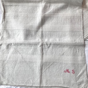 May include: A light beige linen towel with horizontal textured stripes. The towel has a woven pattern and is embroidered with the letters "M S" in red. The towel is folded.