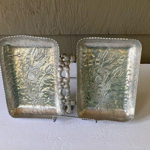May include: A silver-colored, two-section serving tray with a floral design. The tray has a hammered texture and a decorative handle. The floral design is embossed on the tray's surface. The tray is supported by a wire stand.