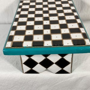 May include: A rectangular wooden riser featuring a black and white checkerboard pattern on the top surface. The edges are painted in a solid teal color. The base displays a black and white diamond pattern, with a notched cutout in the center.