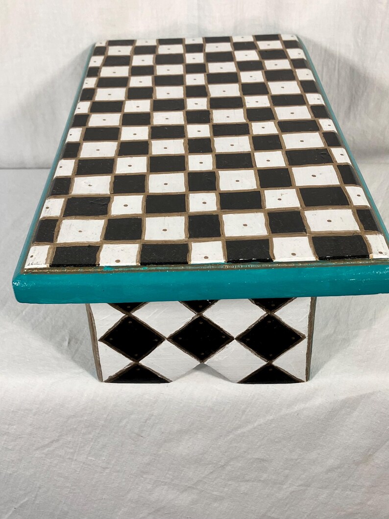May include: A rectangular wooden riser featuring a black and white checkerboard pattern on the top surface. The edges are painted in a solid teal color. The base displays a black and white diamond pattern, with a notched cutout in the center.