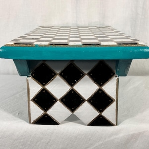 May include: A small wooden stool featuring a flat top with a black and white checkerboard pattern. The base is painted with a black and white diamond pattern. A vibrant teal horizontal trim piece connects the top to the base, adding a pop of color.