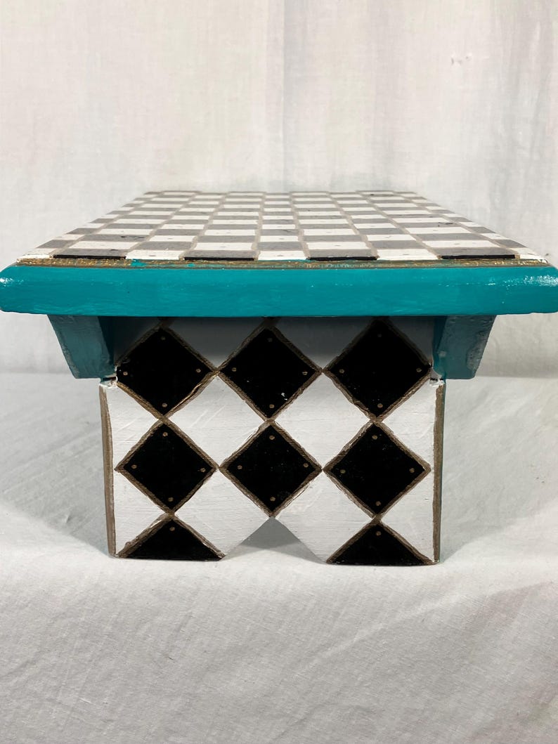 May include: A small wooden stool featuring a flat top with a black and white checkerboard pattern. The base is painted with a black and white diamond pattern. A vibrant teal horizontal trim piece connects the top to the base, adding a pop of color.