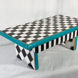 May include: A rectangular wooden stool featuring a black and white checkered top surface. The edges are painted in a bright teal color. The legs display a black and white diamond pattern on the sides and vertical black and white stripes.