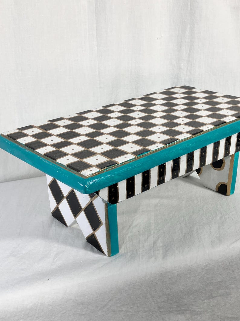 May include: A rectangular wooden stool featuring a black and white checkered top surface. The edges are painted in a bright teal color. The legs display a black and white diamond pattern on the sides and vertical black and white stripes.