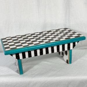 May include: A rectangular wooden step stool featuring a black and white checkered top surface. The side panels are decorated with vertical black and white stripes, while the edges are painted in a vibrant teal color with black polka dots.
