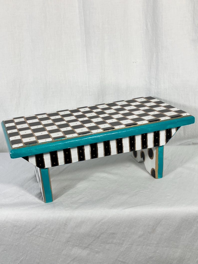 May include: A rectangular wooden step stool featuring a black and white checkered top surface. The side panels are decorated with vertical black and white stripes, while the edges are painted in a vibrant teal color with black polka dots.