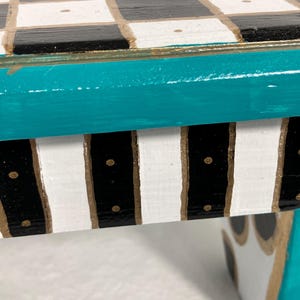 May include: A small table featuring a top surface with a black and white checkered pattern, accented by small gold dots. The table edge is painted in a vibrant teal, while the side apron displays vertical black and white stripes with gold detailing.