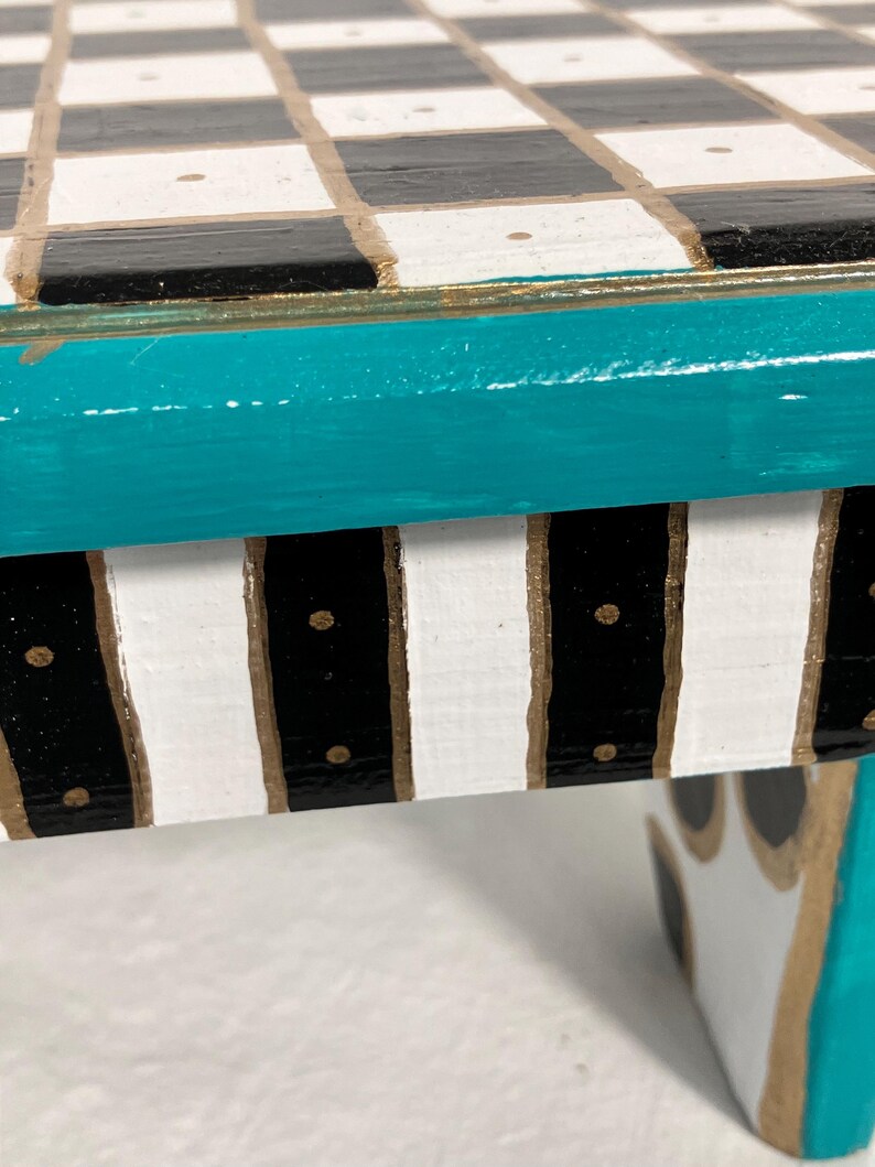 May include: A small table featuring a top surface with a black and white checkered pattern, accented by small gold dots. The table edge is painted in a vibrant teal, while the side apron displays vertical black and white stripes with gold detailing.