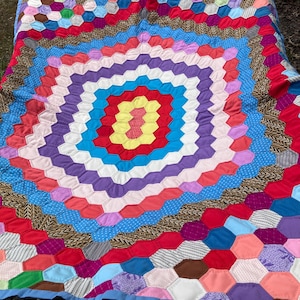 May include: A colorful handmade quilt with a geometric pattern of hexagons in various colors, including red, blue, purple, and yellow. The quilt is displayed on a hammock, showcasing its vibrant design and craftsmanship.
