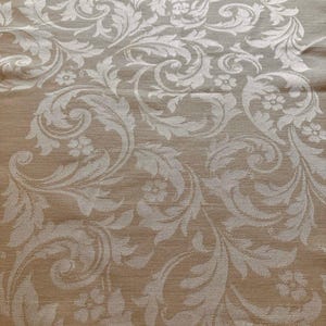 May include: Close-up of a beige fabric with a repeating white floral pattern. The design features stylized leaves and swirling elements, creating a textured surface. The fabric has a subtle sheen, suggesting a luxurious material.