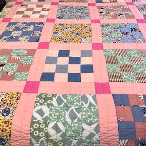 May include: A patchwork quilt with a grid pattern. The quilt features squares of various patterned fabrics in shades of blue, pink, yellow, and green, separated by pink borders. The quilt is likely made of cotton and is a vintage item.