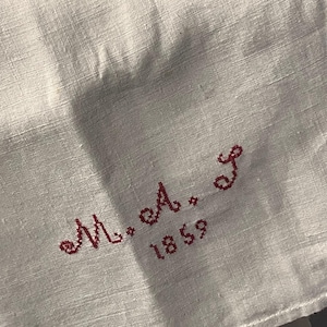May include: Close-up of a white linen handkerchief with red embroidered initials "M. A. J." and the year "1859". The fabric has a subtle texture and is folded, with the embroidery in the corner.
