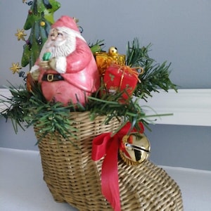May include: A woven wicker Christmas stocking filled with festive decorations. It features a Santa Claus figurine, a small Christmas tree, and wrapped gifts. A red ribbon and a gold bell adorn the stocking, creating a holiday display.