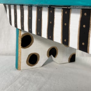 May include: A small wooden riser or stool featuring a vibrant teal top surface. The side panel is decorated with vertical black and white stripes accented by small gold dots. The legs are white with large black polka dots outlined in gold paint.