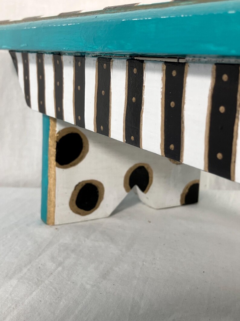 May include: A small wooden riser or stool featuring a vibrant teal top surface. The side panel is decorated with vertical black and white stripes accented by small gold dots. The legs are white with large black polka dots outlined in gold paint.