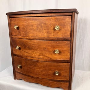 May include: A three-drawer wooden dresser with a curved front and brass knobs. The dresser is stained in a warm, medium-brown color, with a darker top. The drawers have a slight curve, adding a touch of elegance to the design.