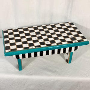 May include: A rectangular wooden step stool featuring a black and white checkered pattern on the top surface. The sides of the stool are painted in a teal color, while the legs display a black and white vertical striped pattern with teal accents.