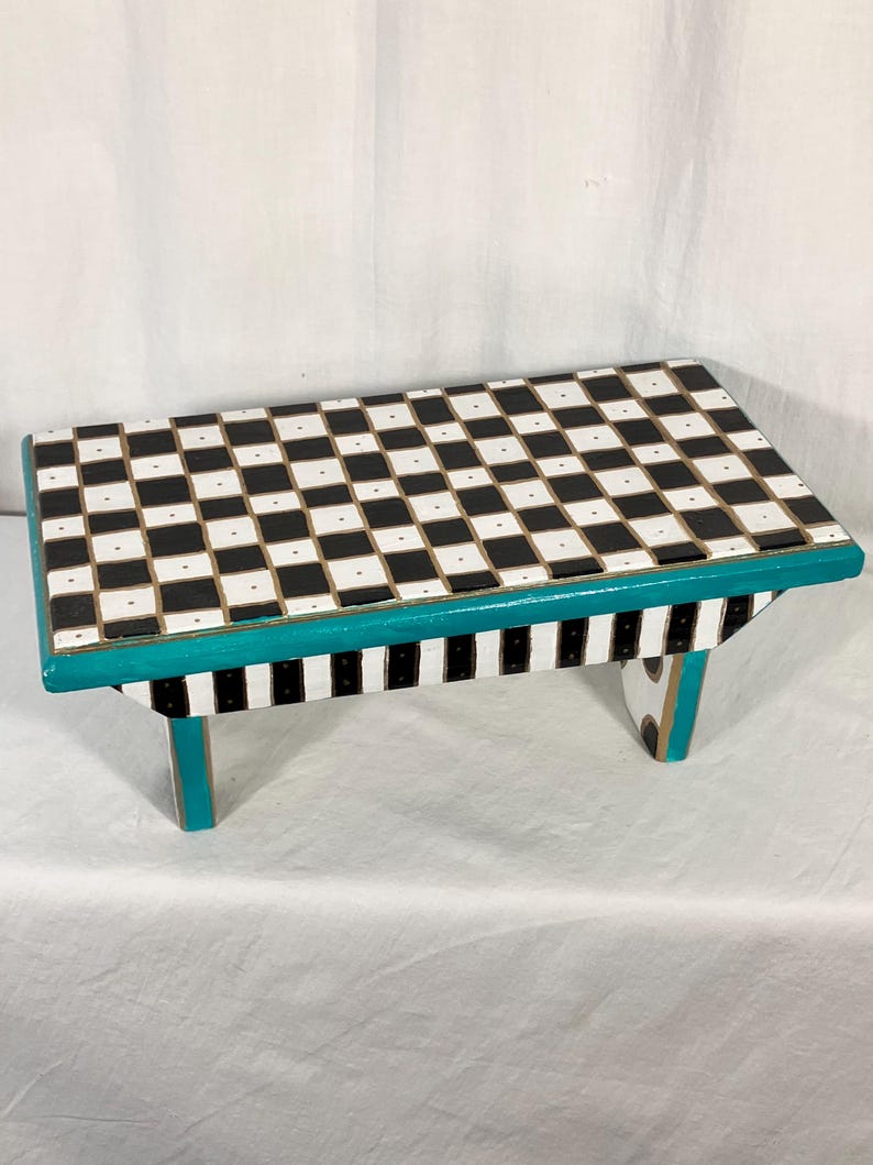 May include: A rectangular wooden step stool featuring a black and white checkered pattern on the top surface. The sides of the stool are painted in a teal color, while the legs display a black and white vertical striped pattern with teal accents.