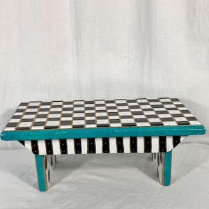 May include: A small wooden stool featuring a black and white checkered top surface. The side frame is painted in a vibrant teal color, while the base support features vertical black and white stripes. The legs are also accented with teal trim.