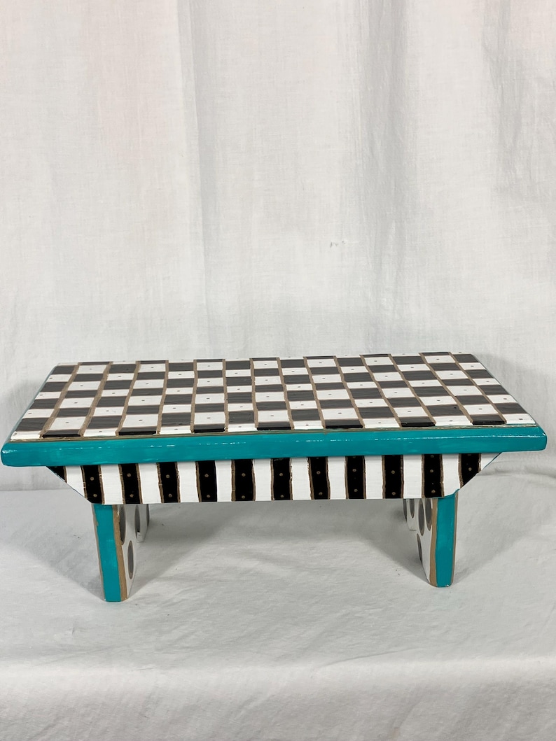 May include: A small wooden stool featuring a black and white checkered top surface. The side frame is painted in a vibrant teal color, while the base support features vertical black and white stripes. The legs are also accented with teal trim.