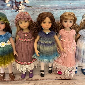 May include: Five dolls wearing knitted dresses and hats. The dresses are in shades of blue, green, pink, and purple. The hats are white, light blue, and green. The dolls have brown, blonde, and curly brown hair.