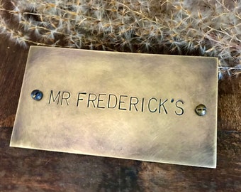 May include: A brass plaque with the text "MR FREDERICK'S" engraved on it. The plaque is attached to a wooden surface with two screws.