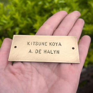 Pode incluir: Uma placa de identificação retangular de latão com o texto "KITSUNE KOYA" acima de "A. DE HALYN". A placa tem dois pequenos orifícios, um em cada extremidade. O fundo é folhagem verde desfocada.