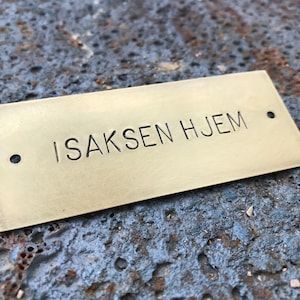 Pu&ograve; includere: Una placca in ottone con il testo "ISAKSEN HJEM" inciso su di essa. La placca ha due fori nella parte superiore e inferiore.